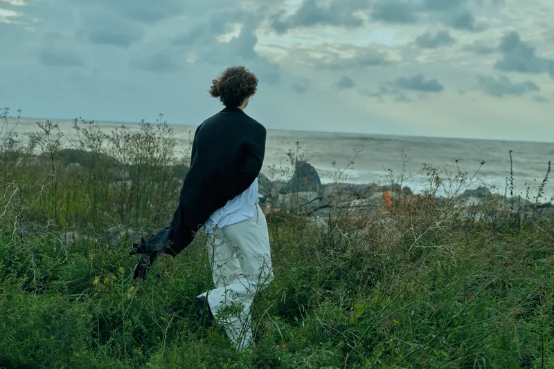 Walker along a windswept shoreline, grasses and rocks around, wearing a dark jacket and light pants, facing the sea beneath a cloudy sky, a quiet coastal scene