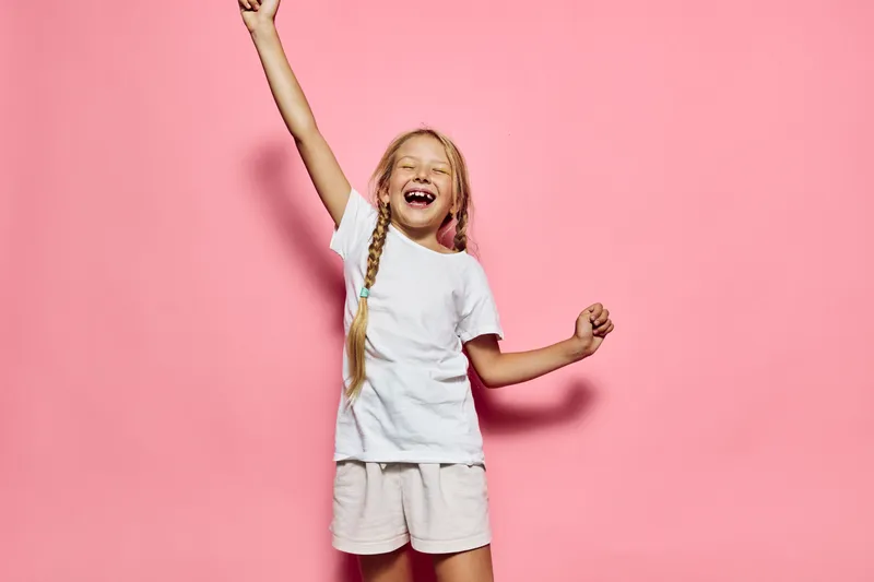 Excited smiling girl with braided hair raises hand wearing white t-shirt and shorts on pink background. Happy expression.