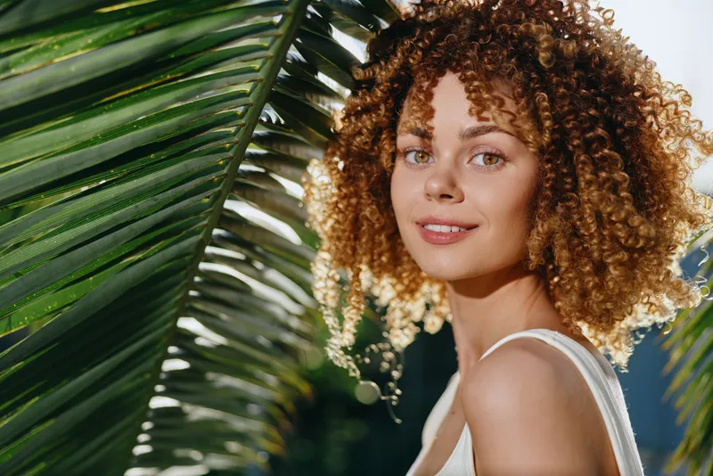 curly woman smiling outdoors with tropical palm leaves, natural light, summer vibe, casual white top, warm and inviting atmosphere
