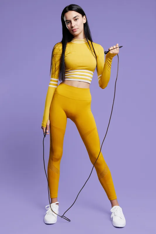 Woman sport fitness wearing yellow activewear, holding a jump rope, standing confidently against a purple background in a full-body portrait with white sneakers.