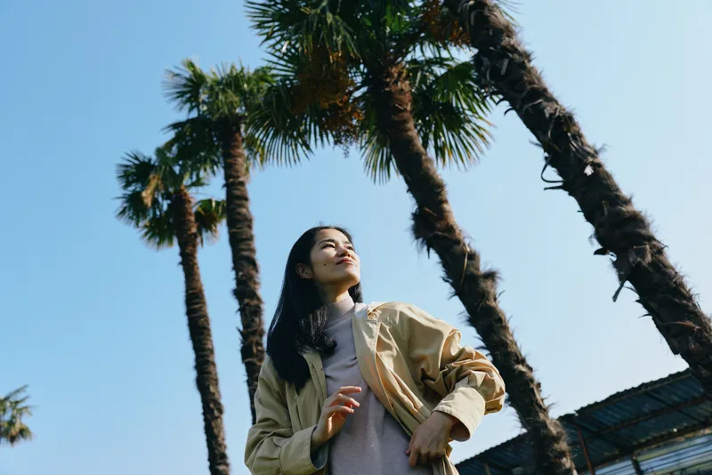 Palm trees frame a woman outdoors in a beige jacket gazing upward under clear sunshine and blue sky showcasing casual fashion and a carefree mood today