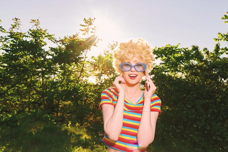 Cheerful woman outdoors in a striped shirt, enjoying a sunlit moment with a retro vibe, oversized sunglasses, and a relaxed pose amid lush greenery