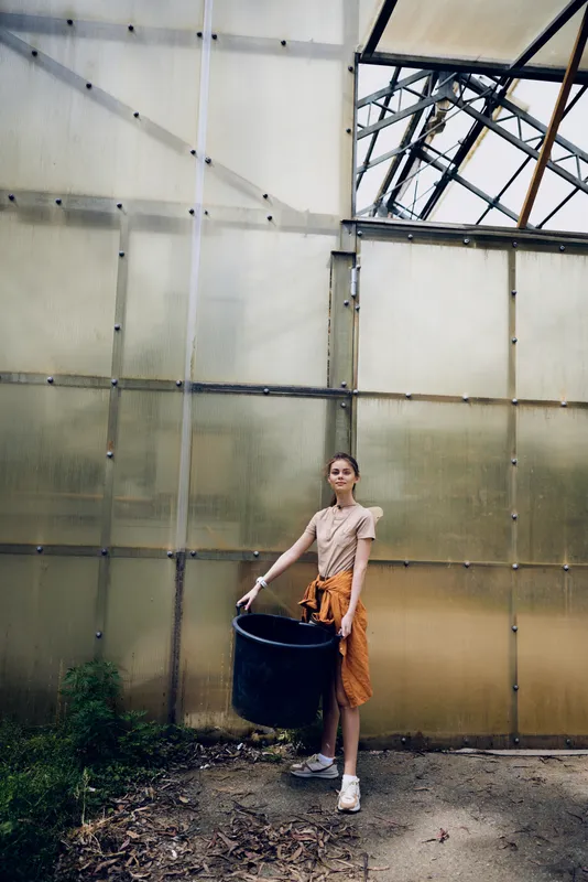 Woman greenhouse standing and holding a large black bucket, gardening in a rustic glass structure, casual outfit with orange sweater tied at waist, natural light and earthy floor detail.