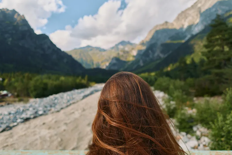 A person stands with back to the camera, facing a majestic valley surrounded by mountains and a flowing river. The scene conveys solitude, exploration, and natural beauty.
