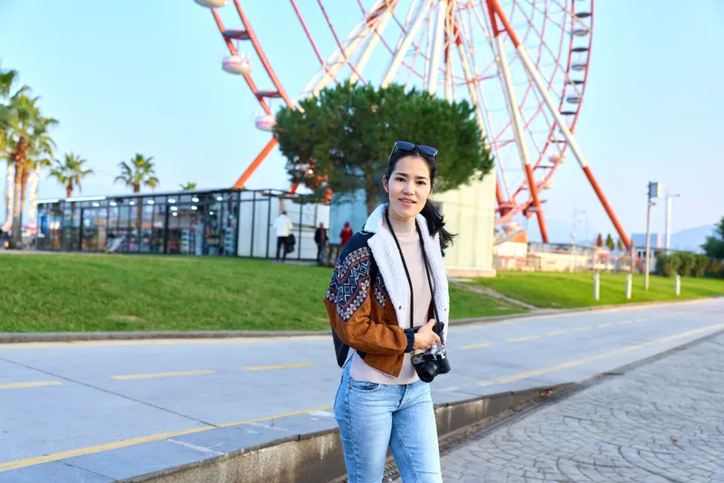 Woman in casual fashion posing outdoors near a large amusement wheel in a sunny park, denim jeans, jacket, camera in hand, relaxed stance, travel mood, leisure outing, urban daylight photography