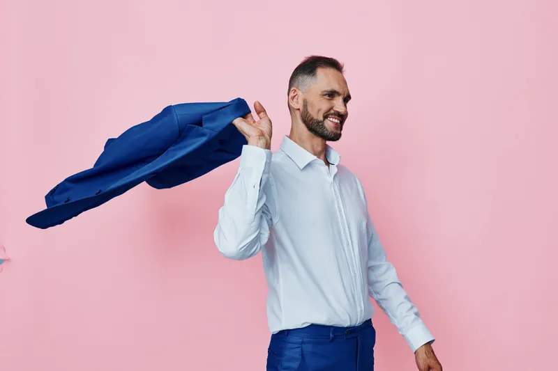 A confident business professional stands against a solid pink background, with a blue cape of fabric over his shoulder, symbolizing triumph and resilience.