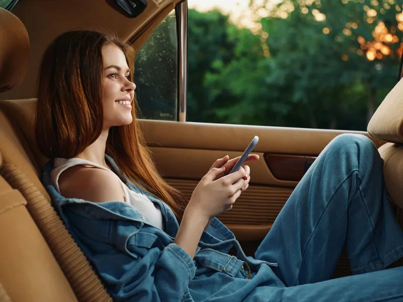 Young woman enjoying her phone in a car, relaxed and smiling, with a casual denim outfit against a green background at sunset.