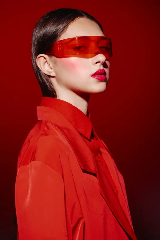 Woman in bold red attire and vibrant makeup wears futuristic red eyewear against a red background. Concept for beauty creativity, fashion, modern style, and artistic portrait.