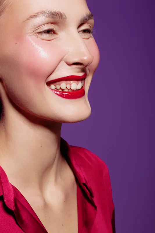 Close-up of smiling woman with red lipstick and shiny skin wearing red blouse against purple background. Concept of beauty, makeup, happiness, and skincare with vibrant colors and natural light.