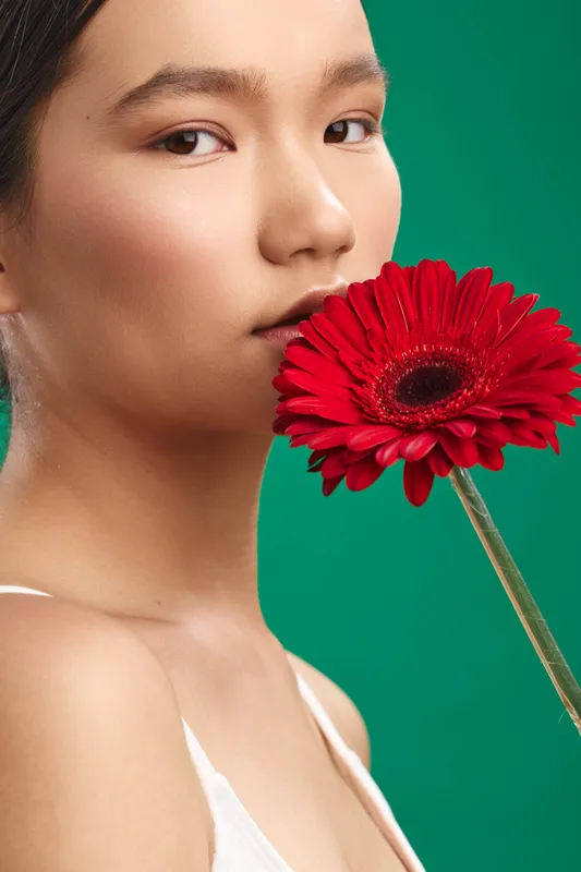 asian woman gazes softly at the camera with a bold red flower held near her lips, set against a vibrant green backdrop, creating a striking beauty portrait with calm, intimate mood and subtle light.