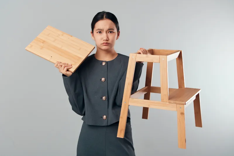 Young woman holding broken wooden chair and detached seat part with unhappy expression in studio. Female in dark outfit facing camera with damaged furniture and frustrated mood.