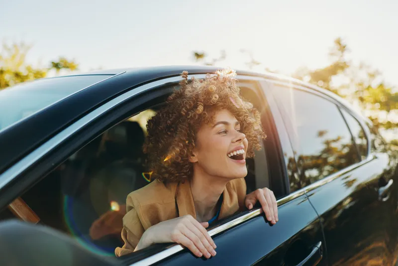 A joyful young adult woman with curly hair leans out of a sunlit car window, laughing as a warm breeze tousles her hair and the landscape rolls by.