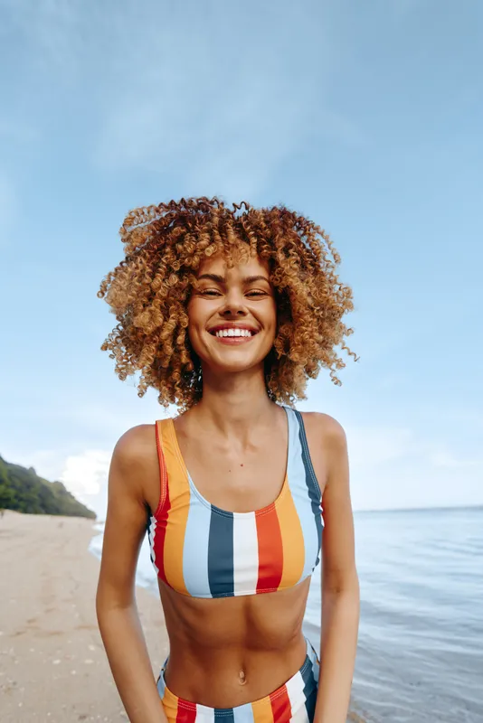 Smiling woman with curly hair wearing colorful striped swimsuit standing on sandy beach near calm sea under clear sky, bright natural light, joyful summer mood