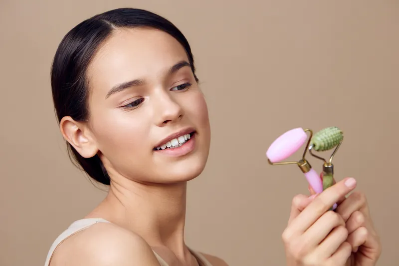 Young woman smiling while holding facial rollers, promoting skincare and self care with pastel colors and a neutral background