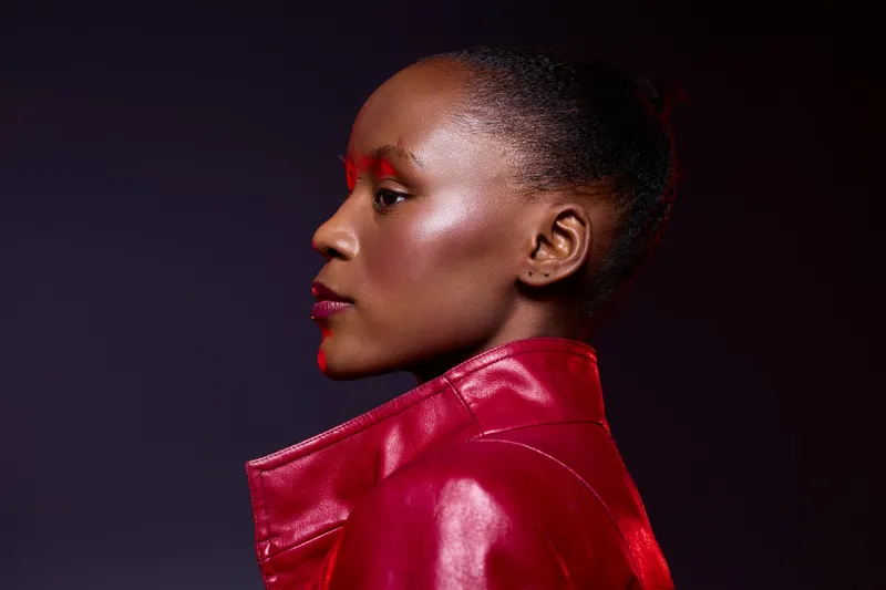 Profile of an African woman with dark skin wearing a red leather jacket against a dark background. The image focuses on fashion, beauty, and elegance with dramatic lighting.