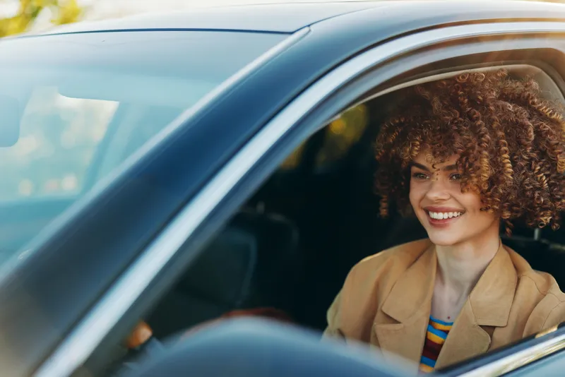 Smiling woman with curly hair sits in a car, leaning toward the window. Natural daylight highlights her cheerful mood and vintage fashion, capturing a warm lifestyle moment of everyday driving.