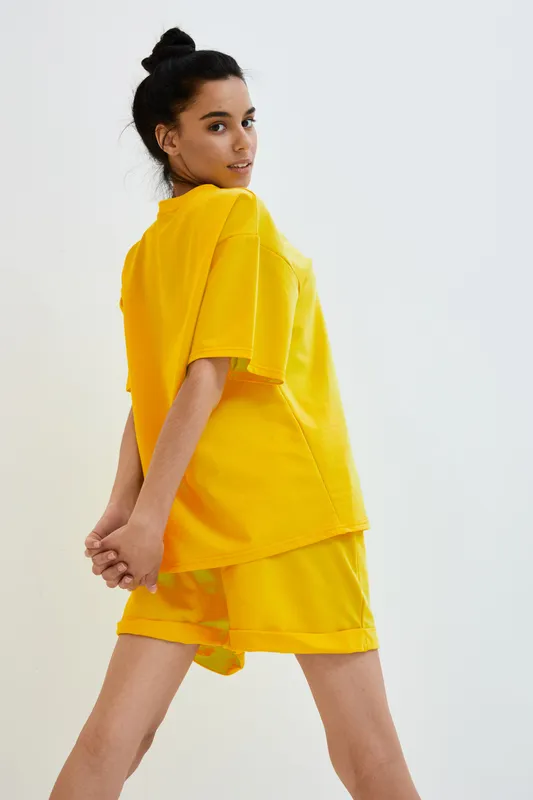young woman in bright yellow casual outfit posing confidently against a plain white background, showcasing modern sporty fashion and a playful expression.