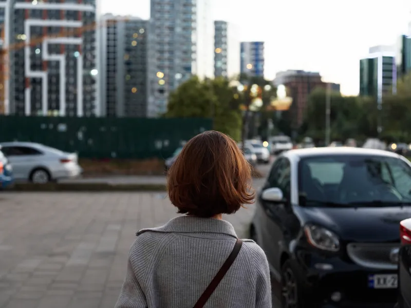 A woman with short brown hair stands with her back to the camera in a modern urban setting, near parked cars and tall buildings.