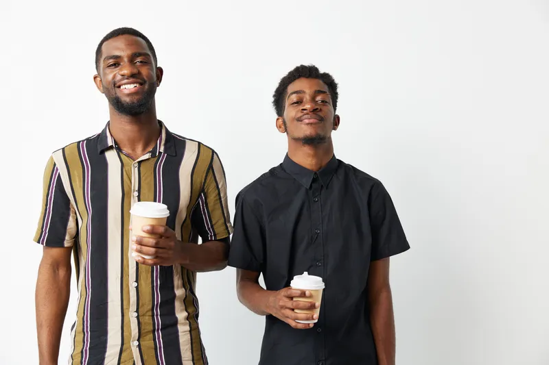 Two smiling African American men standing together with coffee cups in their hands. The stylish outfits add to the vibrant and cheerful atmosphere of the moment.