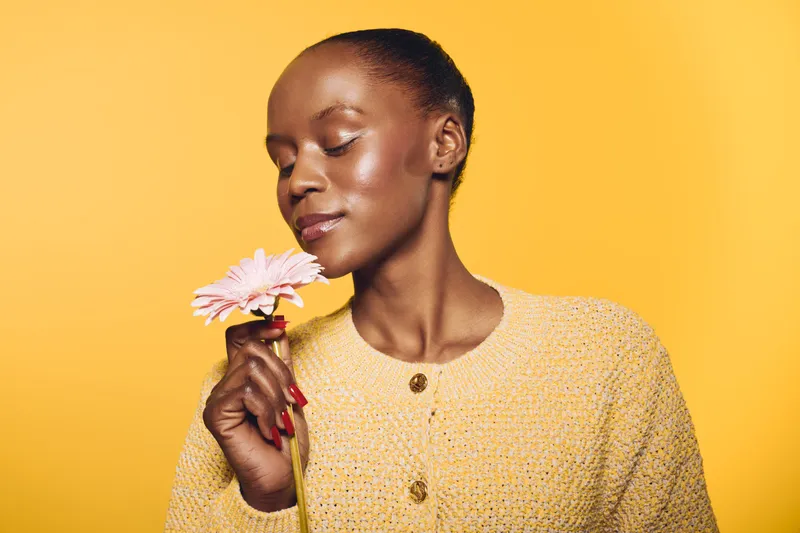 Portrait of a happy African woman with dark skin holding a pink flower close to her face, eyes closed, wearing a yellow knitted sweater on yellow background. Concept of beauty and nature.