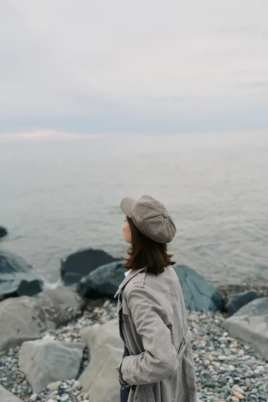 Young woman in a coat and beret stands on rocky shore looking at calm sea under cloudy sky. Concept of solitude, travel, and peaceful nature moments by the water.