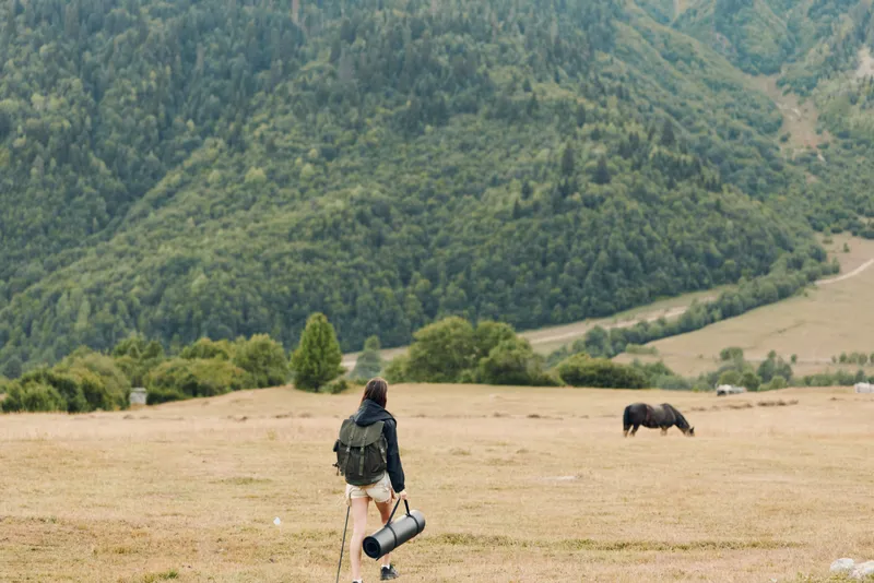A lone traveler with a backpack walks across a wide open field toward distant hills where a calm horse grazes, creating a quiet outdoor scene of exploration, solitude, and natural beauty.