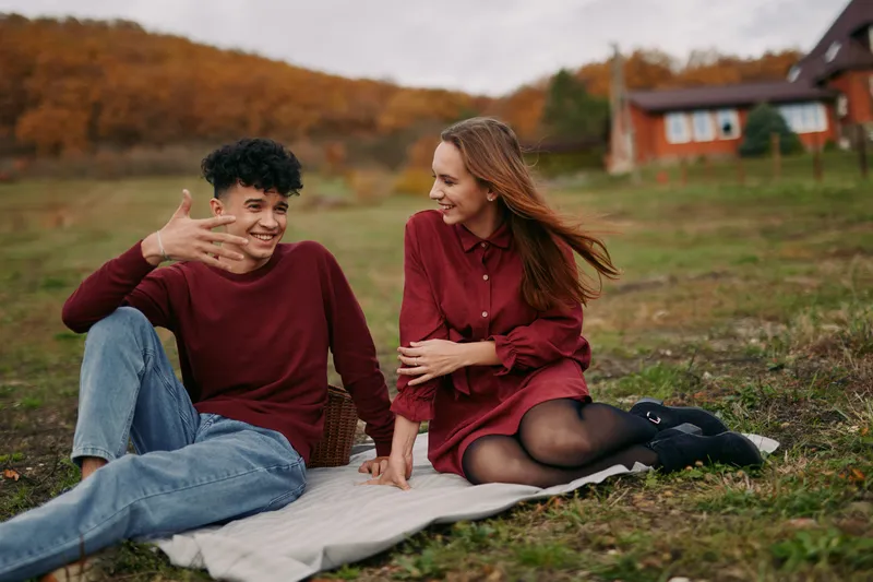 Candid moment of a young couple sharing laughter on a rustic picnic blanket in an autumn meadow, capturing genuine connection, natural expressions and reliable everyday intimacy.