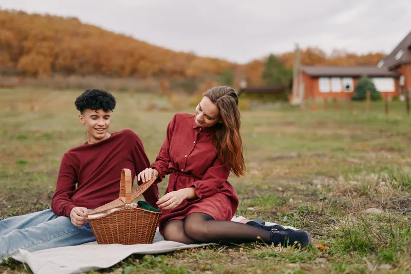 A genuine outdoor picnic scene capturing authentic connection between two young people seated on a blanket, sharing a wicker basket and natural moments of warmth and ease in a rural meadow.