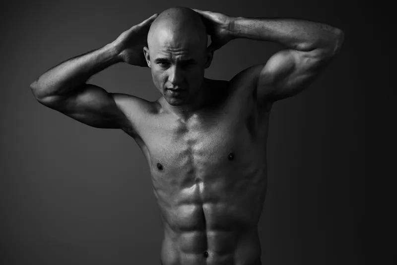 Strong man showcasing his physique, demonstrating confidence and strength in a dramatic black and white portrait, with powerful lighting emphasizing his features.