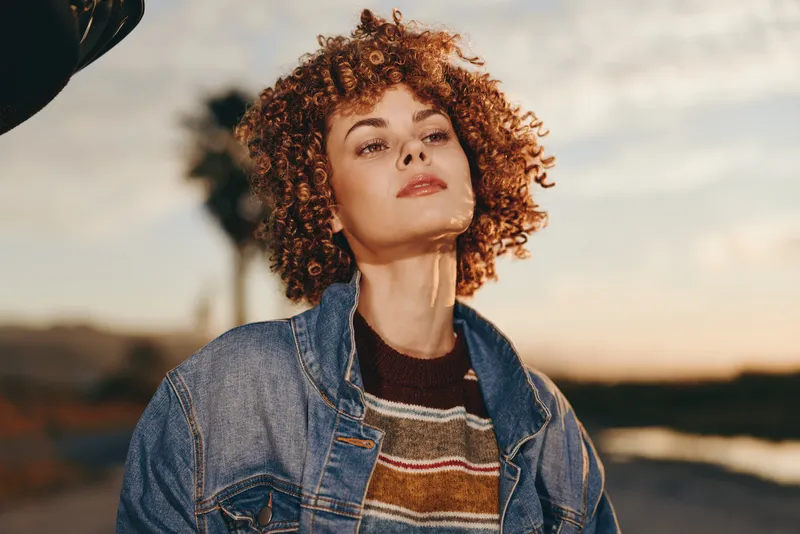 Woman curly hair outdoors sunset denim lifestyle portrait, relaxed expression in soft sunlit breeze, natural tones, evening sky, blurred road and landscape background bokeh style