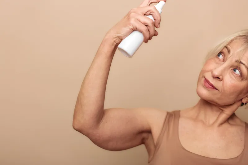 Mature woman with short blonde hair sprays deodorant under her arm, wearing a sleeveless top. Concept for personal hygiene, skincare, and daily self-care routine.