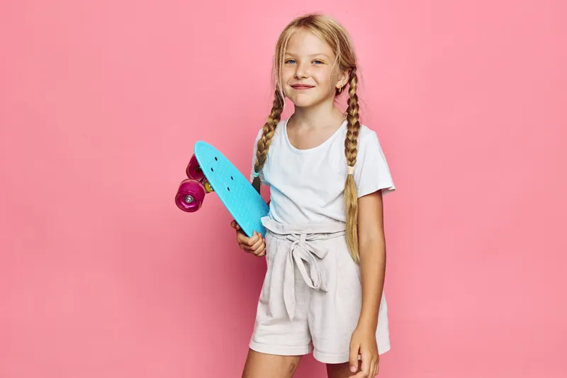 Smiling young girl with blonde hair in braids holds a blue skateboard wearing a white t-shirt and beige shorts. Studio portrait on pink background. Concept for youth and sports.