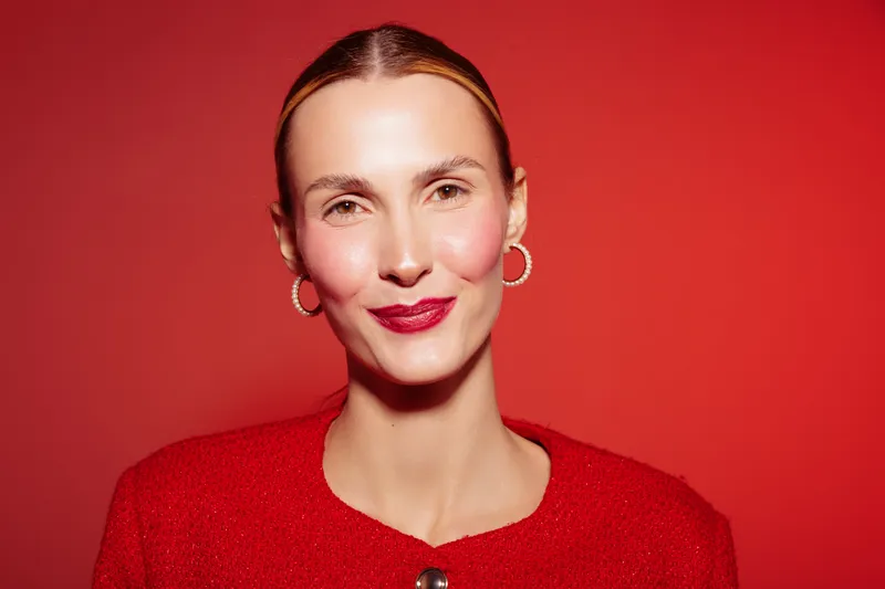 Confident woman with slicked hair and red lipstick smiles against red background wearing red outfit and hoop earrings. Concept of beauty, fashion, and elegance.