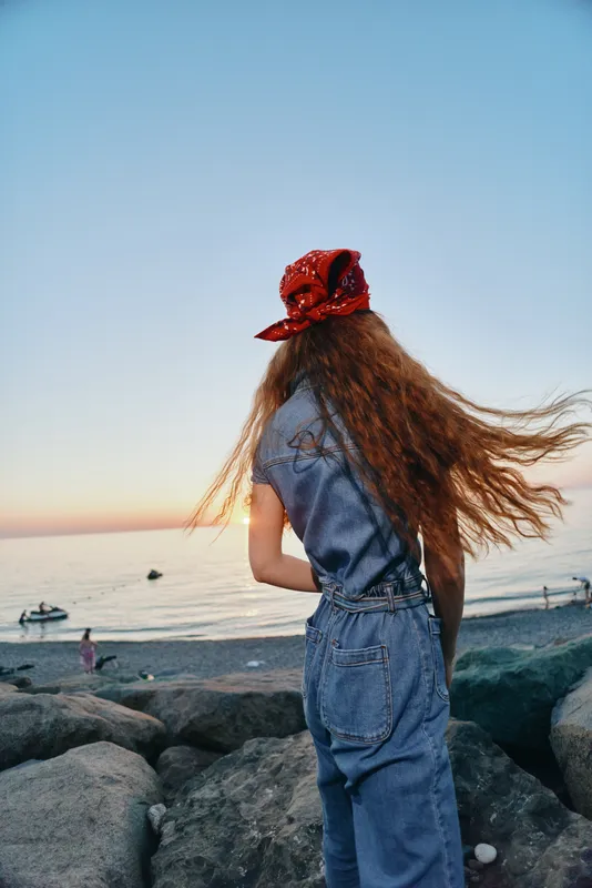 young woman with long hair wearing red bandana and denim outfit standing on rocks by the sea during sunset with clear blue sky