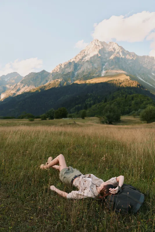 A woman lies on a grassy field near dramatic mountains, resting with a backpack beside her. A moment of pause during outdoor travel, reflecting calm and nature mindfulness.
