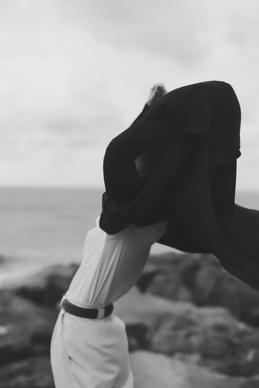 Wind flows through a scarf draped over the head, white outfit on a rocky coast, creating a dramatic, monochrome silhouette, and seashells on shore, mood of resilience and solitude
