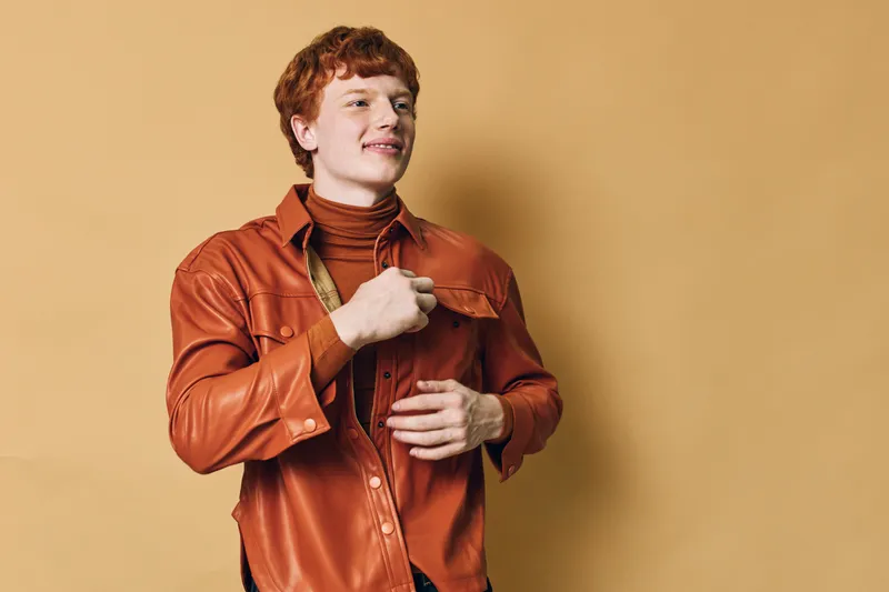 Young man with red hair wearing a brown leather shirt and turtleneck standing against beige background. Casual fashion style with confident expression and relaxed pose.