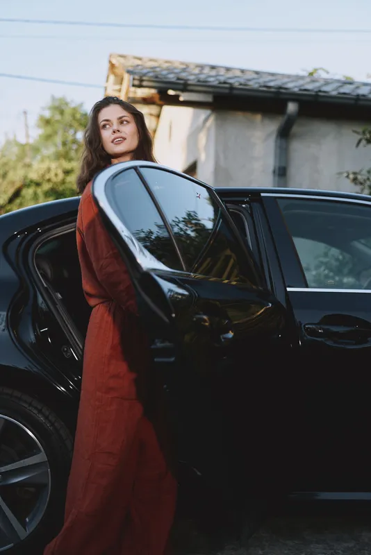 Woman in a rust jumpsuit steps out of a sleek black car in a suburban driveway. Casual lifestyle, confident mood, candid moment, warm daylight lighting, natural background