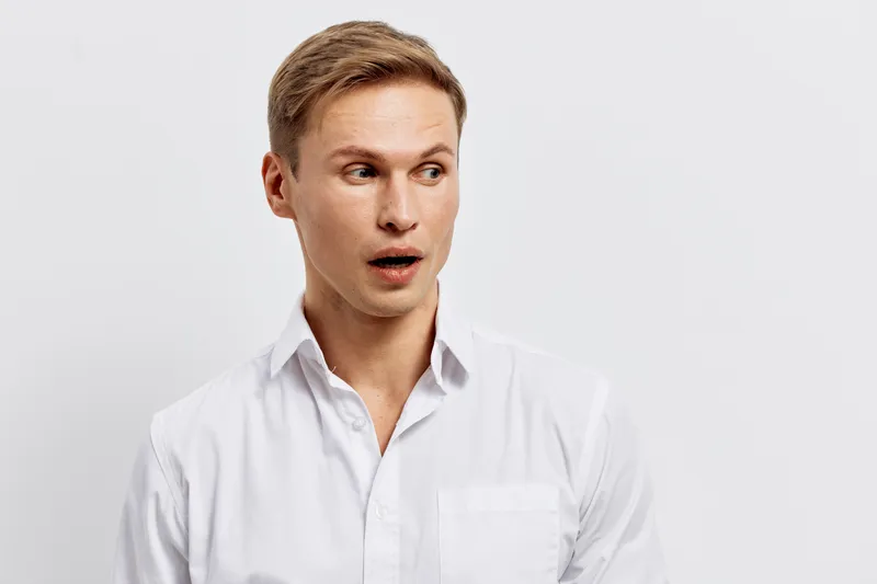 Surprised man with open mouth and wide eyes wearing white shirt looking sideways isolated on plain white background studio portrait. People lifestyle concept