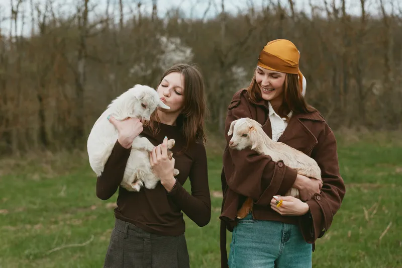Cheerful women outdoors, holding goat kids, warm interaction, soft breeze, natural colors, rural setting, candid smiles, close bond, relaxed posture, sunlit moment, gentle atmosphere