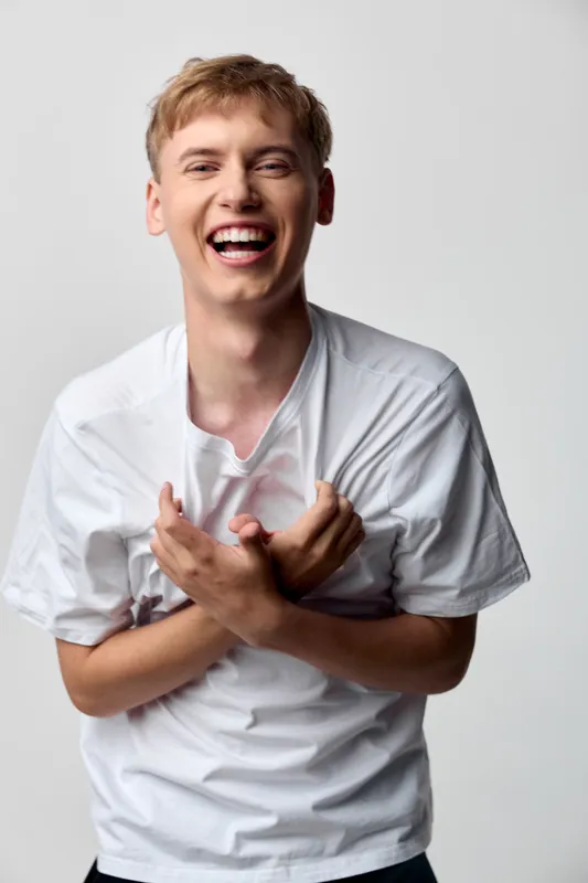 Joyful young man in a white shirt laughing with arms crossed at chest, candid moment of happiness, casual style, bright studio setting, energetic and upbeat mood, and positive energy too