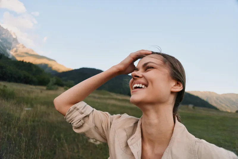 A woman enjoying the outdoors, smiling in a sunny landscape. An open field and distant mountains convey a carefree lifestyle and a strong connection with nature.