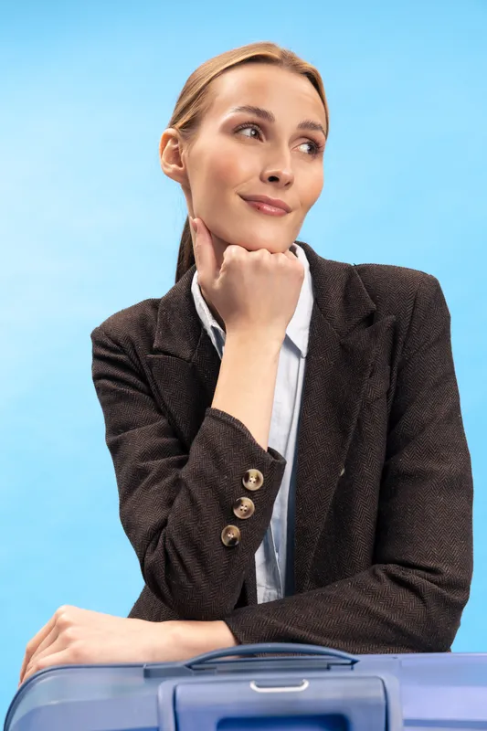 Thoughtful young woman in business attire rests chin on hand, sitting behind suitcase against blue background. Concept of travel planning, business trip, and professional journey.