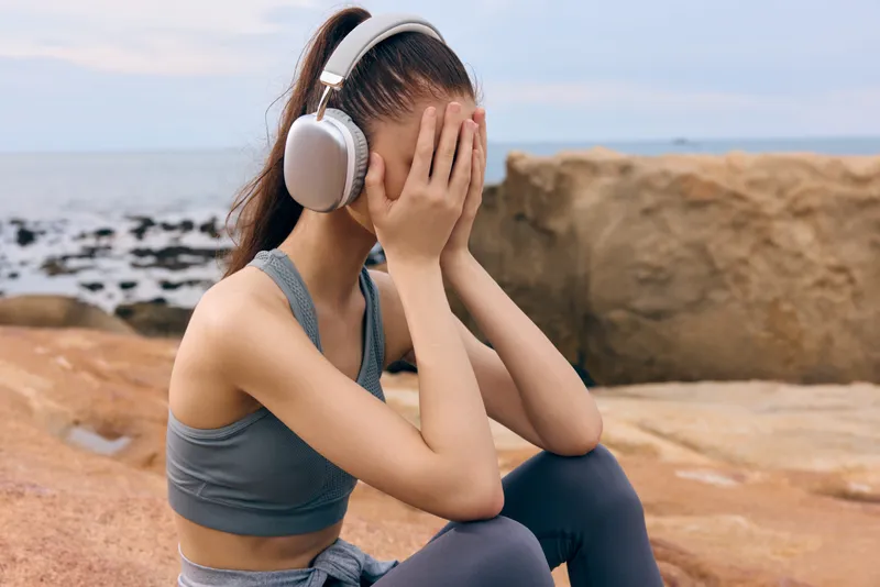 Young woman in sportswear sitting on rocky beach, feeling emotional with hands on face, headphones on, sea background, overcast sky, conveying stress and solitude