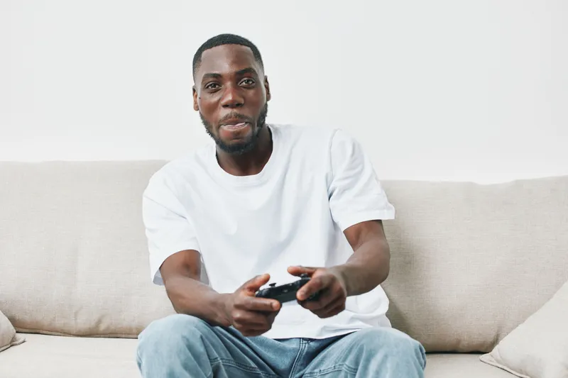 Young African man enjoying video games on a couch, wearing a casual white t shirt and smiling with excitement, showcasing a relaxed and enjoyable atmosphere