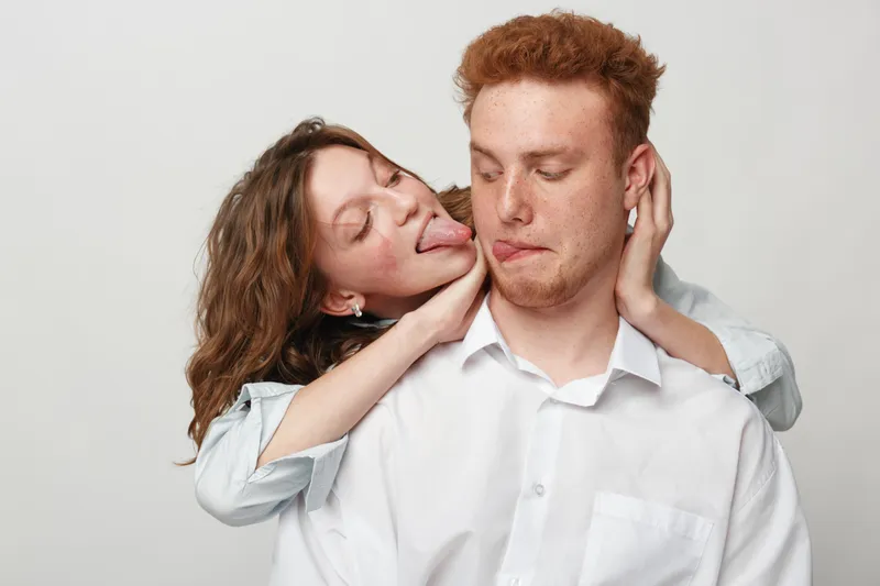 Playful young woman with curly hair teasing her redheaded boyfriend by sticking out her tongue while hugging him from behind. Casual couple interaction on white background. Concept of fun and