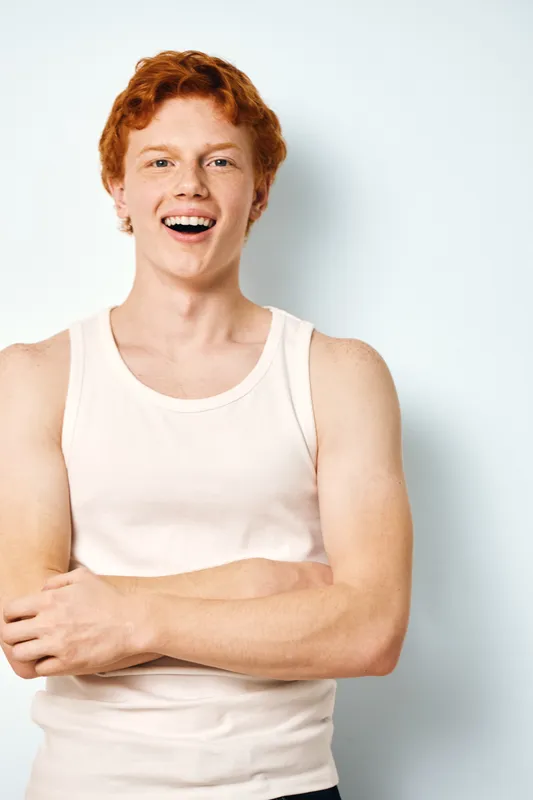 Young redhead man smiling with arms crossed wearing white sleeveless shirt standing against white plain background. Casual confident male portrait with natural expression and relaxed posture.