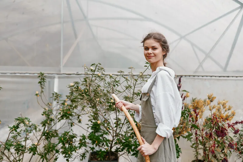 Young woman in a greenhouse, wearing a light sweater and overalls, smiling as she holds a gardening tool among green plants in a bright, airy environment