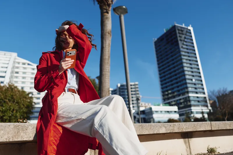 A young woman in a red coat and white pants sits on a city ledge, looking at her phone as a bright blue sky and modern buildings frame a calm, authentic urban moment.