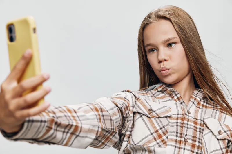 Teenage girl taking selfie with smartphone, wearing plaid shirt, posing with pout lips and long hair on plain light background for social media content.